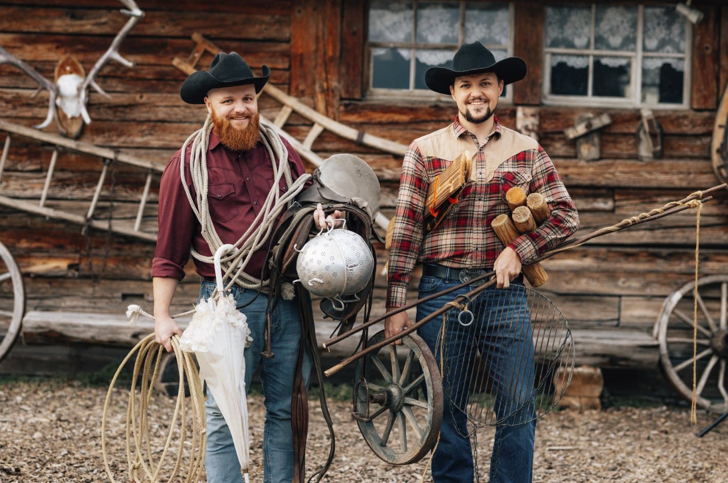 Zwei Cowboys mit Lasso, Milchkanne und Holzscheiten vor dem Blockhaus