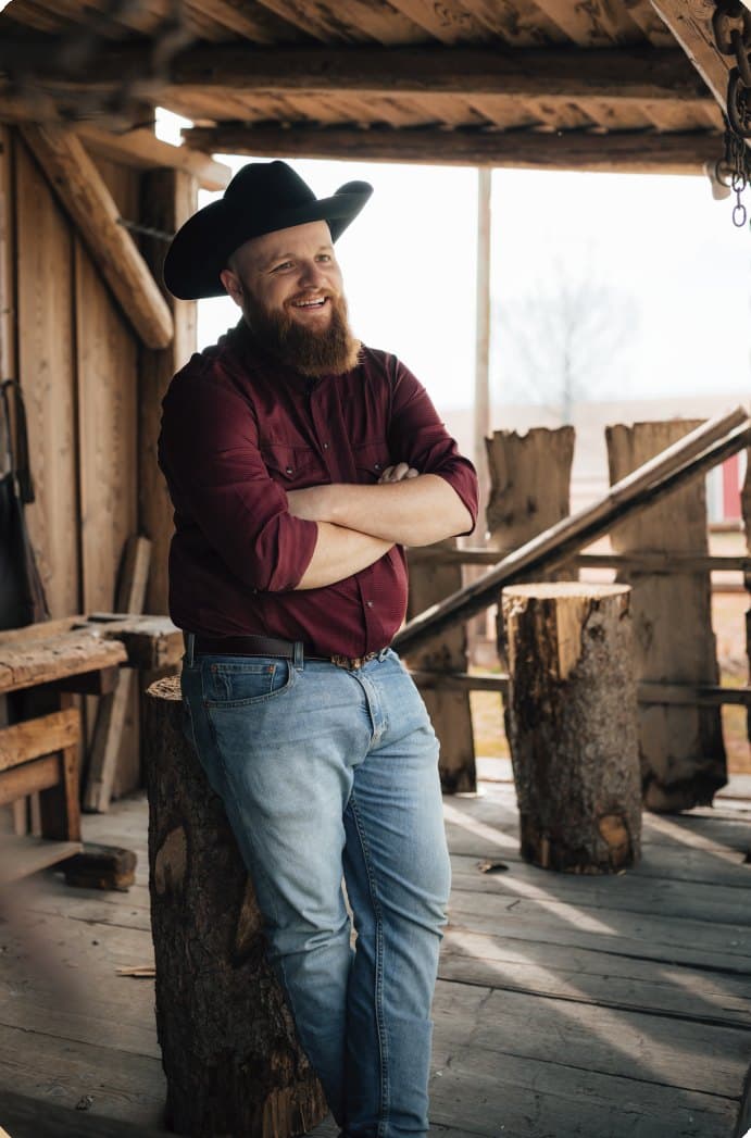 Pascal Luder mit schwarzem Cowboyhut und dunkelrotem Hemd, an einen Holzpfosten gelehnt