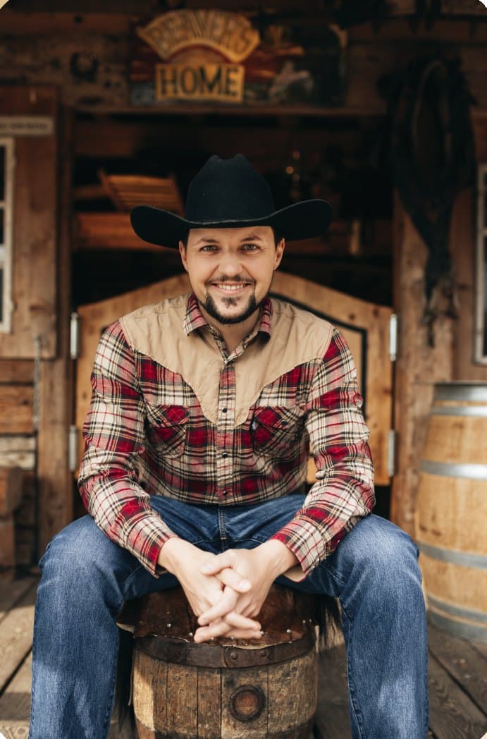 Philipp Luder mit Cowboyhut und Karohemd, auf einem Holzfass vor dem Saloon-Eingang sitzend